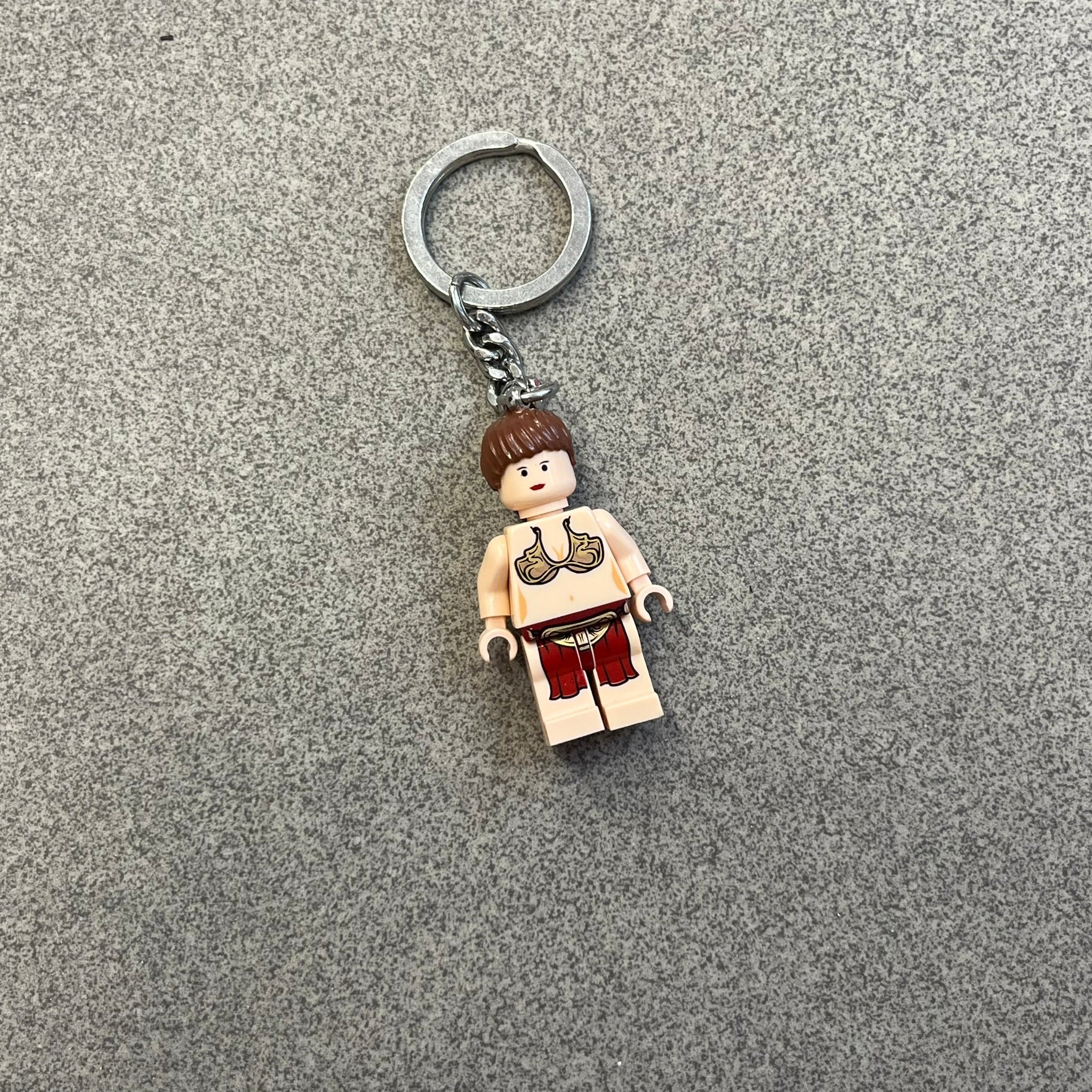 LEGO Star Wars 851938 Princess Leia Key Chain is a 2007 LEGO Gear accessory featuring a Princess Leia minifigure attached to a metal key ring and chain. The figure is permanently fixed to the chain and cannot be removed. The minifigure depicts Princess Leia in her bikini outfit from Jabba’s sail barge scene in Star Wars: Return of the Jedi.