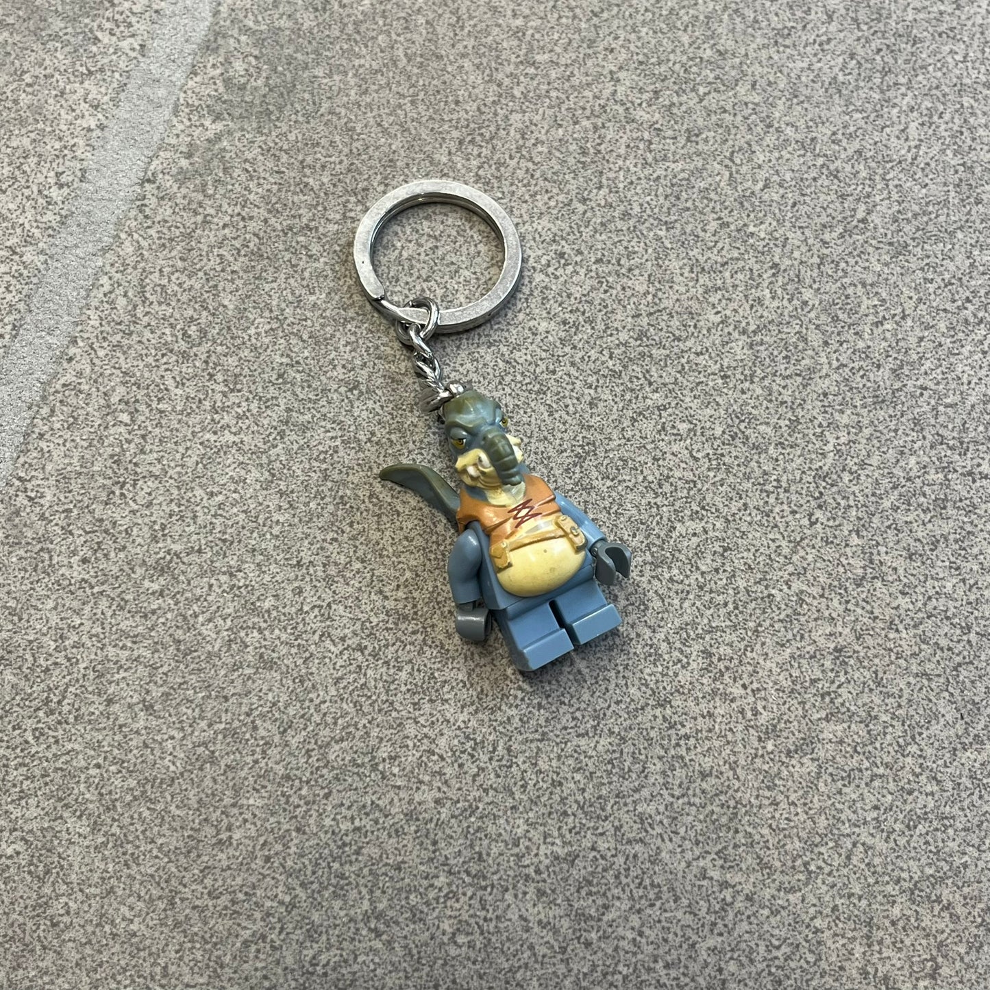 LEGO Star Wars 853413 Watto Key Chain is a 2012 LEGO Gear accessory featuring an authentic LEGO Watto minifigure attached to a metal key ring and chain. The Watto figure is permanently fixed to the chain and cannot be removed. The design represents Watto, the Toydarian junk dealer character from Star Wars: The Phantom Menace.