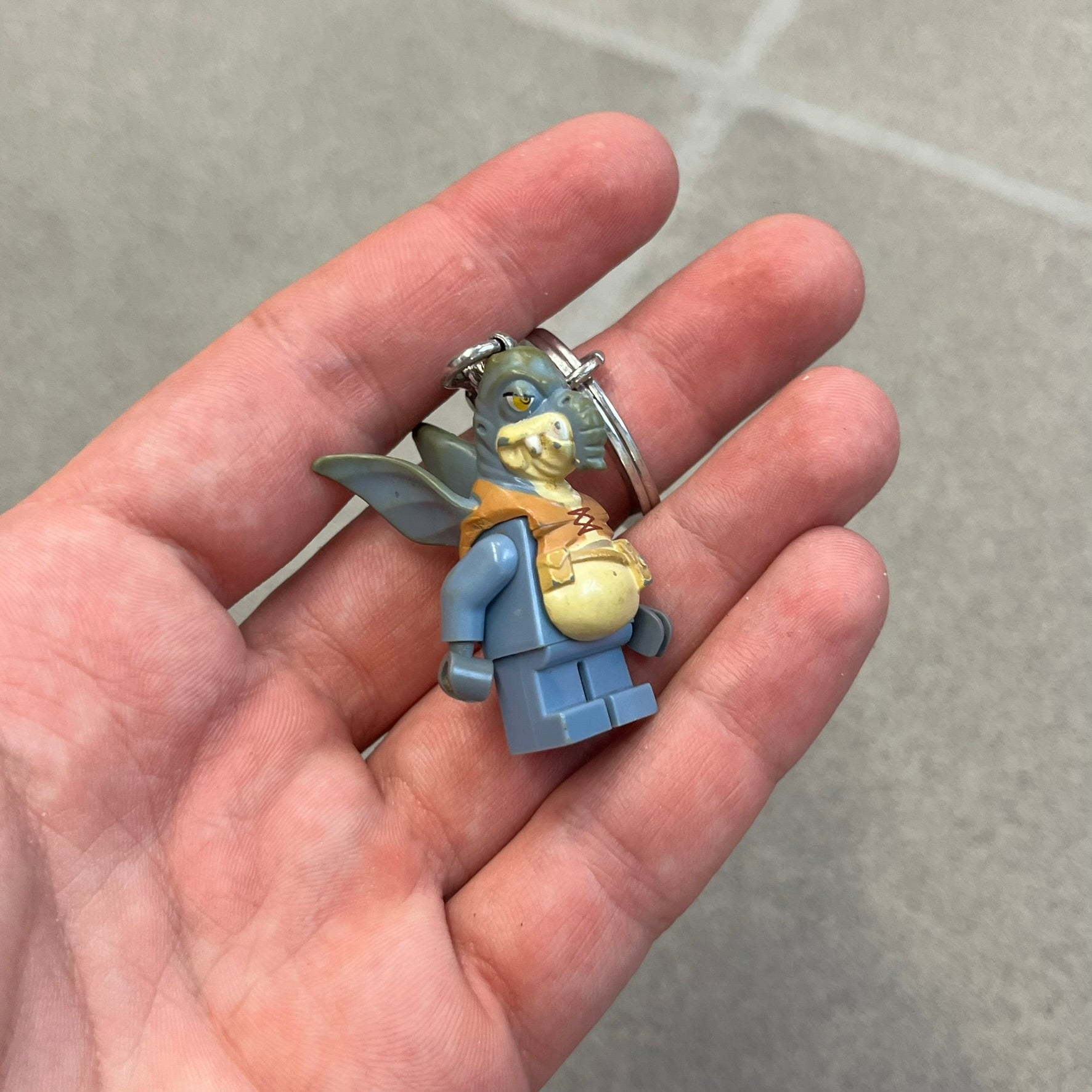 LEGO Star Wars 853413 Watto Key Chain is a 2012 LEGO Gear accessory featuring an authentic LEGO Watto minifigure attached to a metal key ring and chain. The Watto figure is permanently fixed to the chain and cannot be removed. The design represents Watto, the Toydarian junk dealer character from Star Wars: The Phantom Menace.