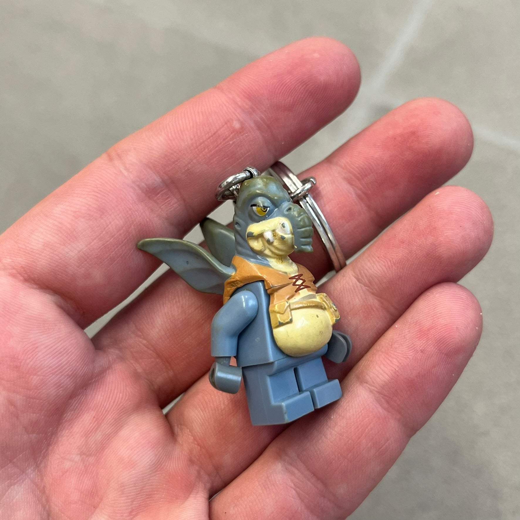 LEGO Star Wars 853413 Watto Key Chain is a 2012 LEGO Gear accessory featuring an authentic LEGO Watto minifigure attached to a metal key ring and chain. The Watto figure is permanently fixed to the chain and cannot be removed. The design represents Watto, the Toydarian junk dealer character from Star Wars: The Phantom Menace.