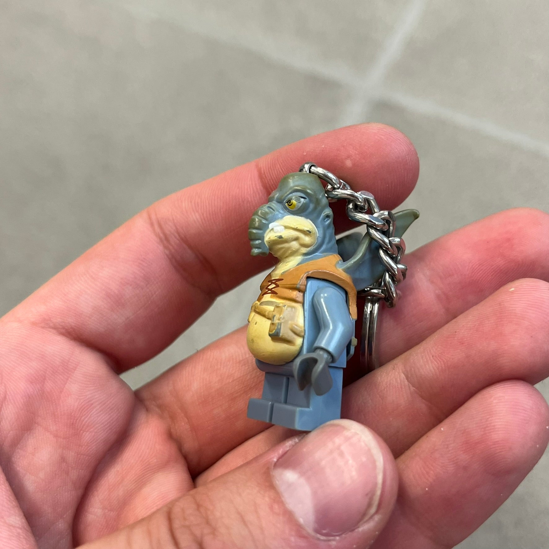LEGO Star Wars 853413 Watto Key Chain is a 2012 LEGO Gear accessory featuring an authentic LEGO Watto minifigure attached to a metal key ring and chain. The Watto figure is permanently fixed to the chain and cannot be removed. The design represents Watto, the Toydarian junk dealer character from Star Wars: The Phantom Menace.