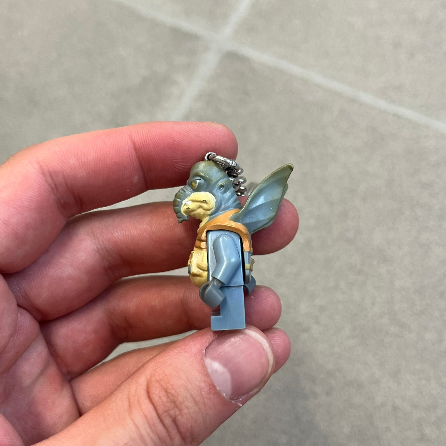 LEGO Star Wars 853413 Watto Key Chain is a 2012 LEGO Gear accessory featuring an authentic LEGO Watto minifigure attached to a metal key ring and chain. The Watto figure is permanently fixed to the chain and cannot be removed. The design represents Watto, the Toydarian junk dealer character from Star Wars: The Phantom Menace.