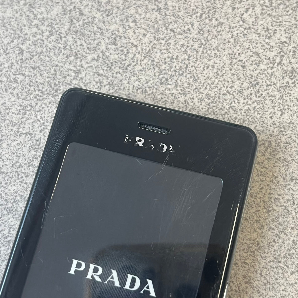LG Prada KE850 Dummy Phone is a promotional display piece replicating the LG Prada KE850, the world's first capacitive touchscreen phone released through a collaboration between LG and Prada. It features the same black case, silver buttons, and top logo as the original, with a fake screen printed with the Prada logo, matching the real phone's size and design.