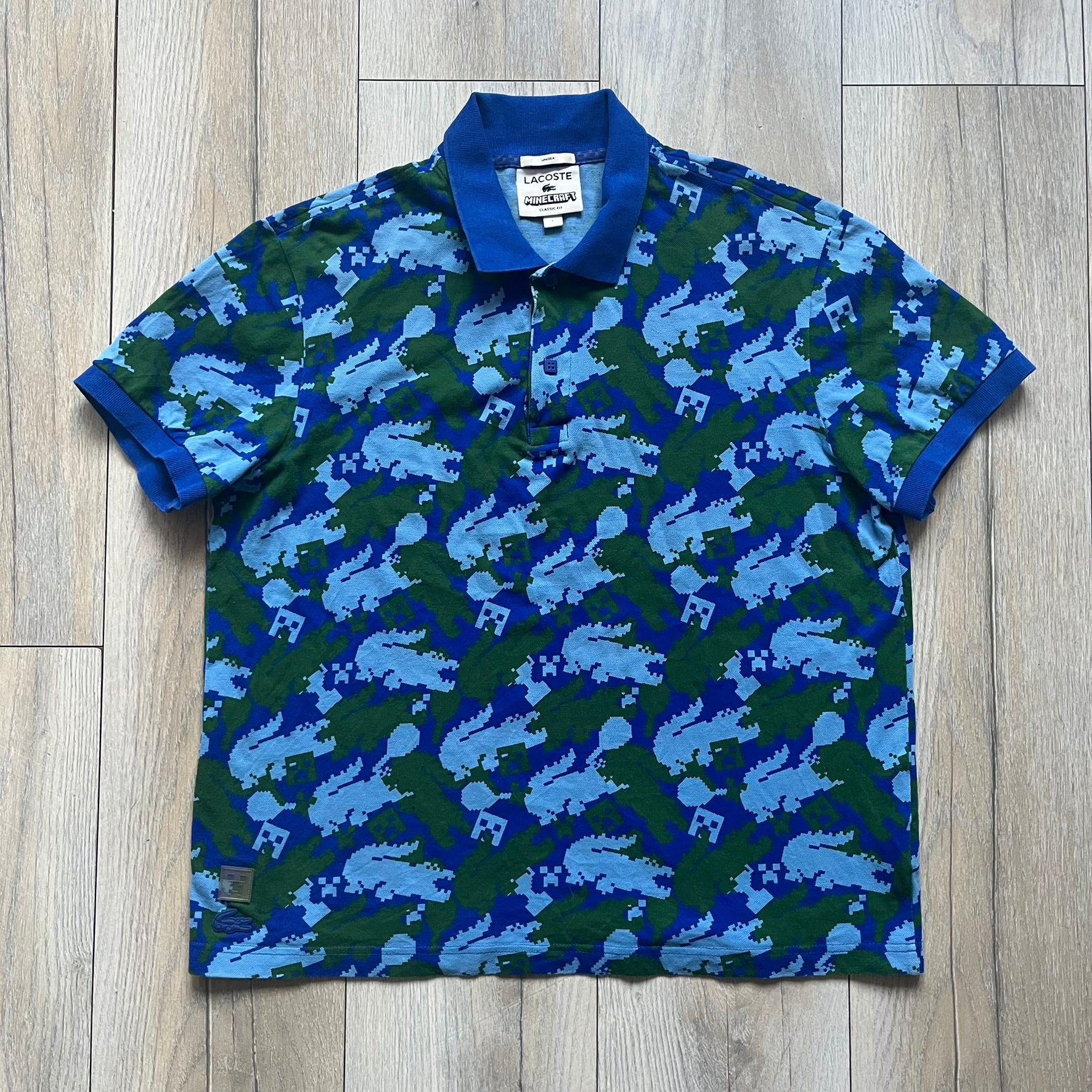 Blue short-sleeve polo shirt from the Lacoste x Minecraft collaboration, released as part of the Spring/Summer 2022 collection (drop in mid-March 2022). Features an all-over print of pixelated Lacoste crocodile logos in green and light blue, interspersed with Minecraft Creeper faces in matching tones. Designed with a solid ribbed polo collar, ribbed sleeve cuffs, and a two-button placket with square buttons featuring Minecraft branding. Finished with a Minecraft badge and a silicone Lacoste crocodile logo p