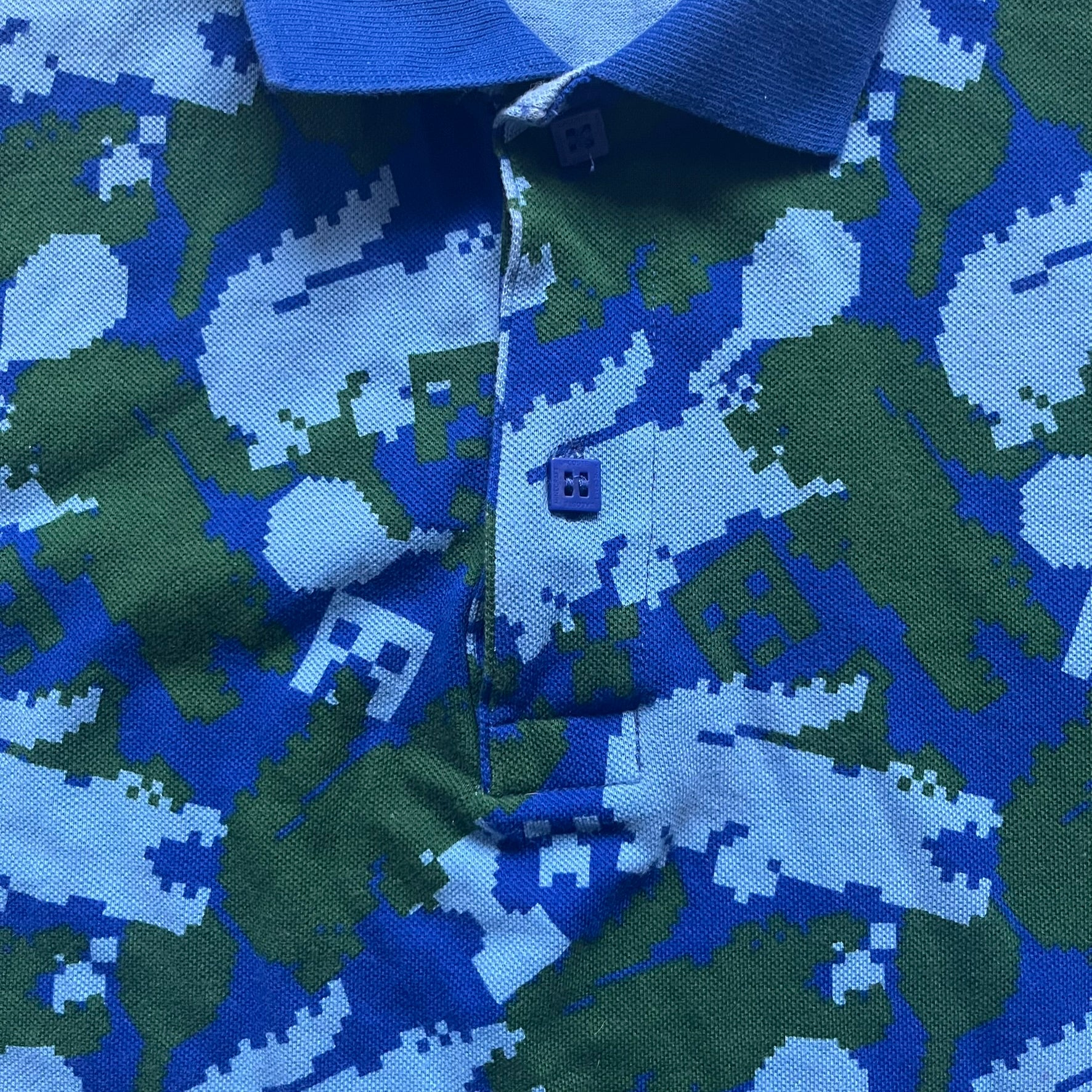 Blue short-sleeve polo shirt from the Lacoste x Minecraft collaboration, released as part of the Spring/Summer 2022 collection (drop in mid-March 2022). Features an all-over print of pixelated Lacoste crocodile logos in green and light blue, interspersed with Minecraft Creeper faces in matching tones. Designed with a solid ribbed polo collar, ribbed sleeve cuffs, and a two-button placket with square buttons featuring Minecraft branding. Finished with a Minecraft badge and a silicone Lacoste crocodile logo p