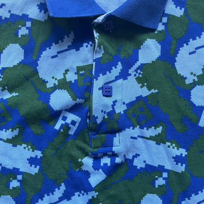 Blue short-sleeve polo shirt from the Lacoste x Minecraft collaboration, released as part of the Spring/Summer 2022 collection (drop in mid-March 2022). Features an all-over print of pixelated Lacoste crocodile logos in green and light blue, interspersed with Minecraft Creeper faces in matching tones. Designed with a solid ribbed polo collar, ribbed sleeve cuffs, and a two-button placket with square buttons featuring Minecraft branding. Finished with a Minecraft badge and a silicone Lacoste crocodile logo p