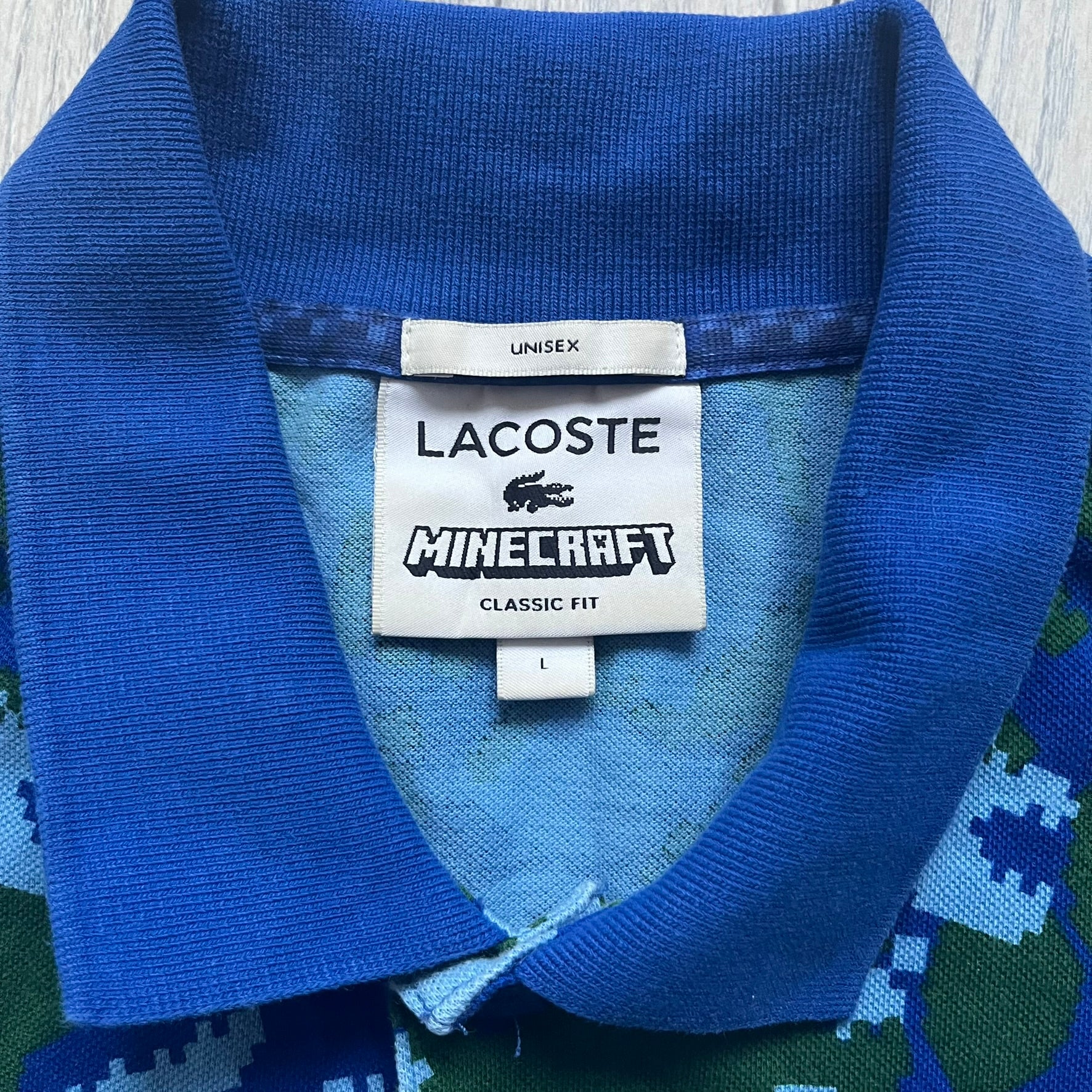 Blue short-sleeve polo shirt from the Lacoste x Minecraft collaboration, released as part of the Spring/Summer 2022 collection (drop in mid-March 2022). Features an all-over print of pixelated Lacoste crocodile logos in green and light blue, interspersed with Minecraft Creeper faces in matching tones. Designed with a solid ribbed polo collar, ribbed sleeve cuffs, and a two-button placket with square buttons featuring Minecraft branding. Finished with a Minecraft badge and a silicone Lacoste crocodile logo p