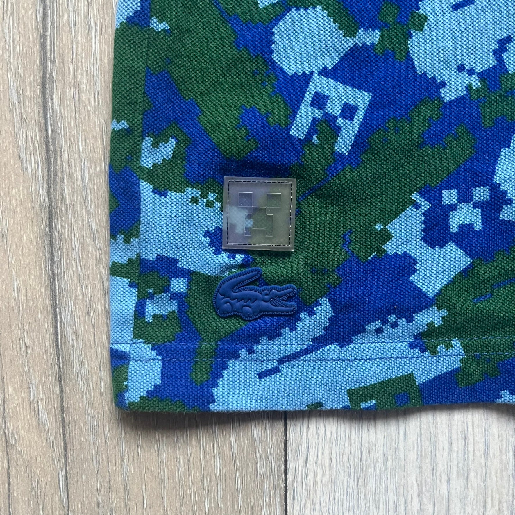 Blue short-sleeve polo shirt from the Lacoste x Minecraft collaboration, released as part of the Spring/Summer 2022 collection (drop in mid-March 2022). Features an all-over print of pixelated Lacoste crocodile logos in green and light blue, interspersed with Minecraft Creeper faces in matching tones. Designed with a solid ribbed polo collar, ribbed sleeve cuffs, and a two-button placket with square buttons featuring Minecraft branding. Finished with a Minecraft badge and a silicone Lacoste crocodile logo p