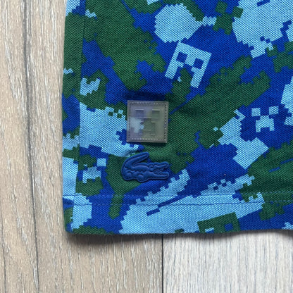 Blue short-sleeve polo shirt from the Lacoste x Minecraft collaboration, released as part of the Spring/Summer 2022 collection (drop in mid-March 2022). Features an all-over print of pixelated Lacoste crocodile logos in green and light blue, interspersed with Minecraft Creeper faces in matching tones. Designed with a solid ribbed polo collar, ribbed sleeve cuffs, and a two-button placket with square buttons featuring Minecraft branding. Finished with a Minecraft badge and a silicone Lacoste crocodile logo p