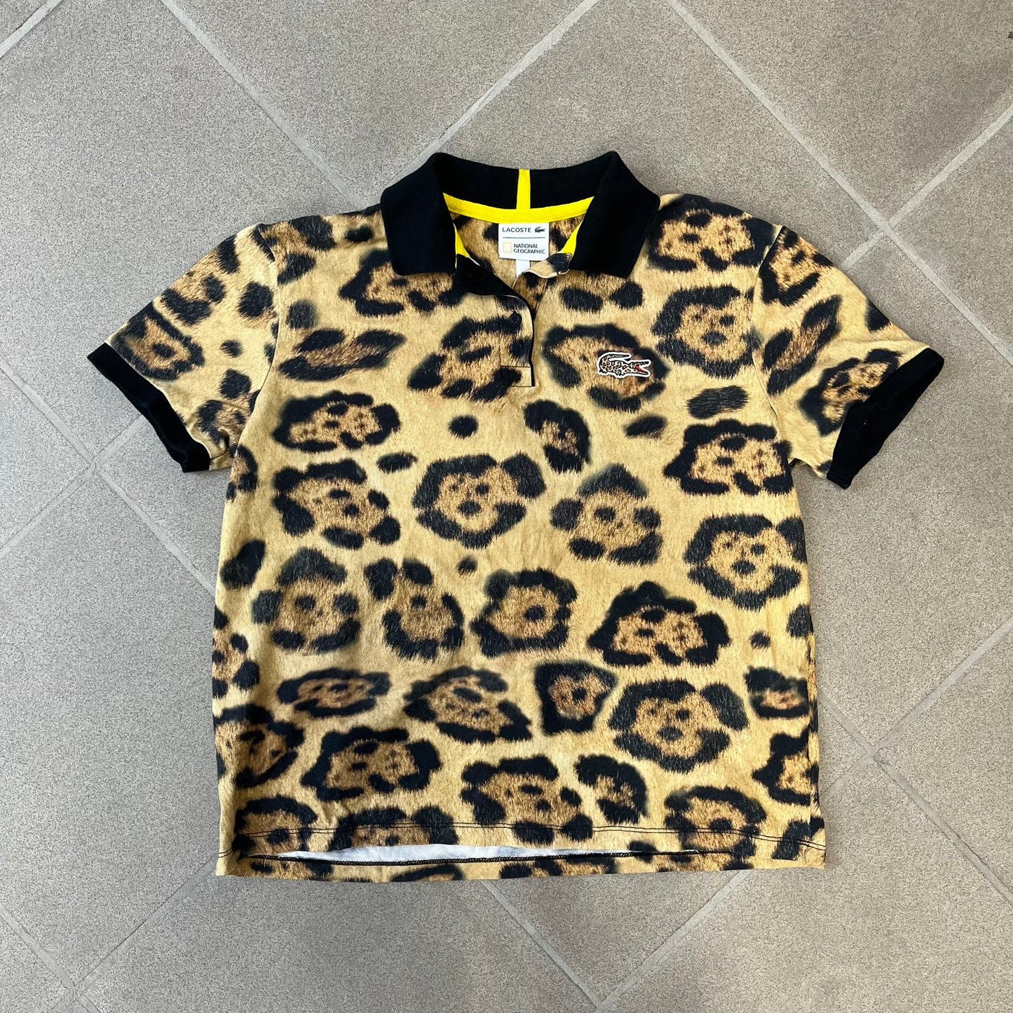 Lacoste x National Geographic AW20 Leopard Polo features an all-over beige, brown and black leopard print pattern across the fabric. The polo has black collar and sleeve hems, with a chest patch featuring the Lacoste crocodile logo in matching pattern colors.