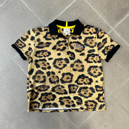 Lacoste x National Geographic AW20 Leopard Polo features an all-over beige, brown and black leopard print pattern across the fabric. The polo has black collar and sleeve hems, with a chest patch featuring the Lacoste crocodile logo in matching pattern colors.