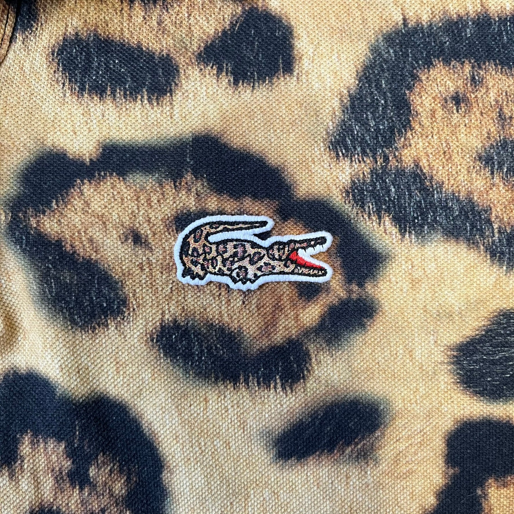 Lacoste x National Geographic AW20 Leopard Polo features an all-over beige, brown and black leopard print pattern across the fabric. The polo has black collar and sleeve hems, with a chest patch featuring the Lacoste crocodile logo in matching pattern colors.
