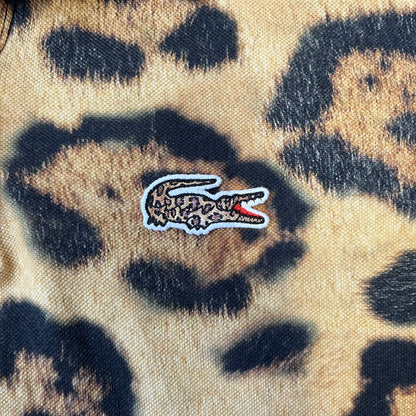 Lacoste x National Geographic AW20 Leopard Polo features an all-over beige, brown and black leopard print pattern across the fabric. The polo has black collar and sleeve hems, with a chest patch featuring the Lacoste crocodile logo in matching pattern colors.