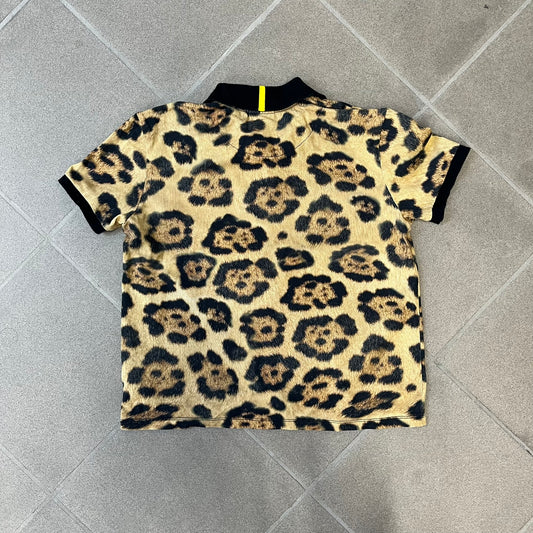 Lacoste x National Geographic AW20 Leopard Polo features an all-over beige, brown and black leopard print pattern across the fabric. The polo has black collar and sleeve hems, with a chest patch featuring the Lacoste crocodile logo in matching pattern colors.