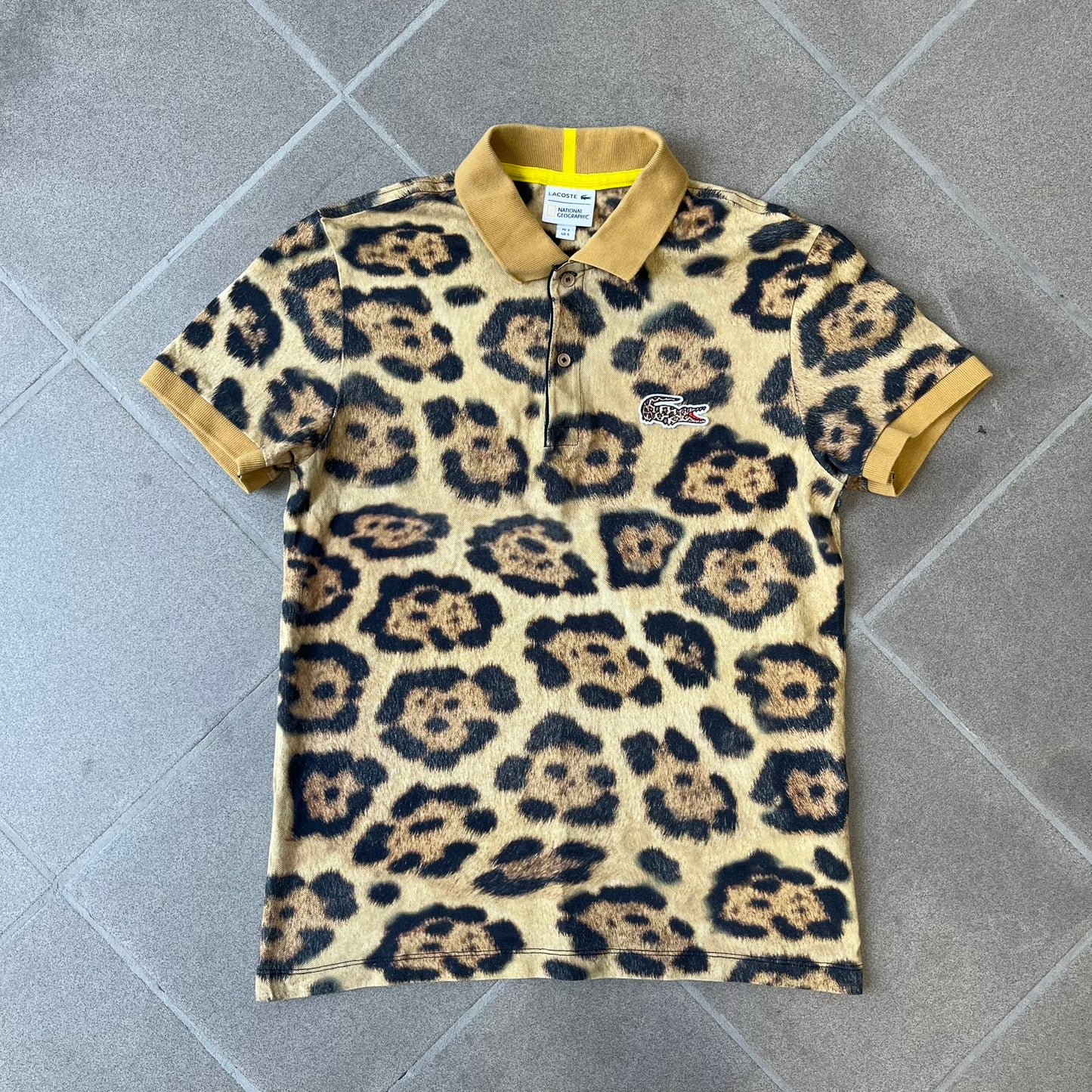 Lacoste x National Geographic AW20 Leopard Polo features an all-over beige, brown and black leopard print pattern across the fabric. The polo has beige collar and sleeve hems, with a chest patch featuring the Lacoste crocodile logo in matching pattern colors.