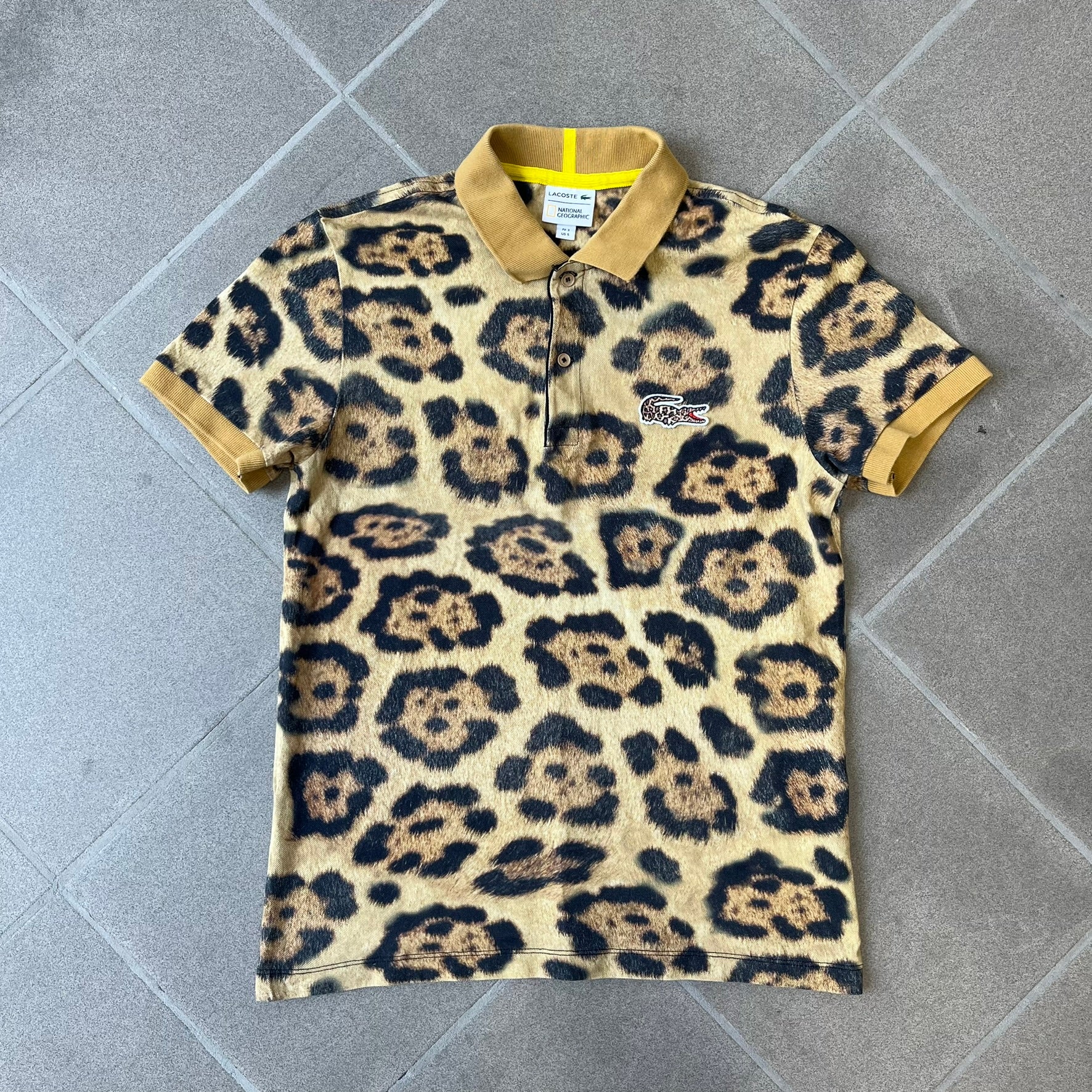 Lacoste x National Geographic AW20 Leopard Polo features an all-over beige, brown and black leopard print pattern across the fabric. The polo has beige collar and sleeve hems, with a chest patch featuring the Lacoste crocodile logo in matching pattern colors.