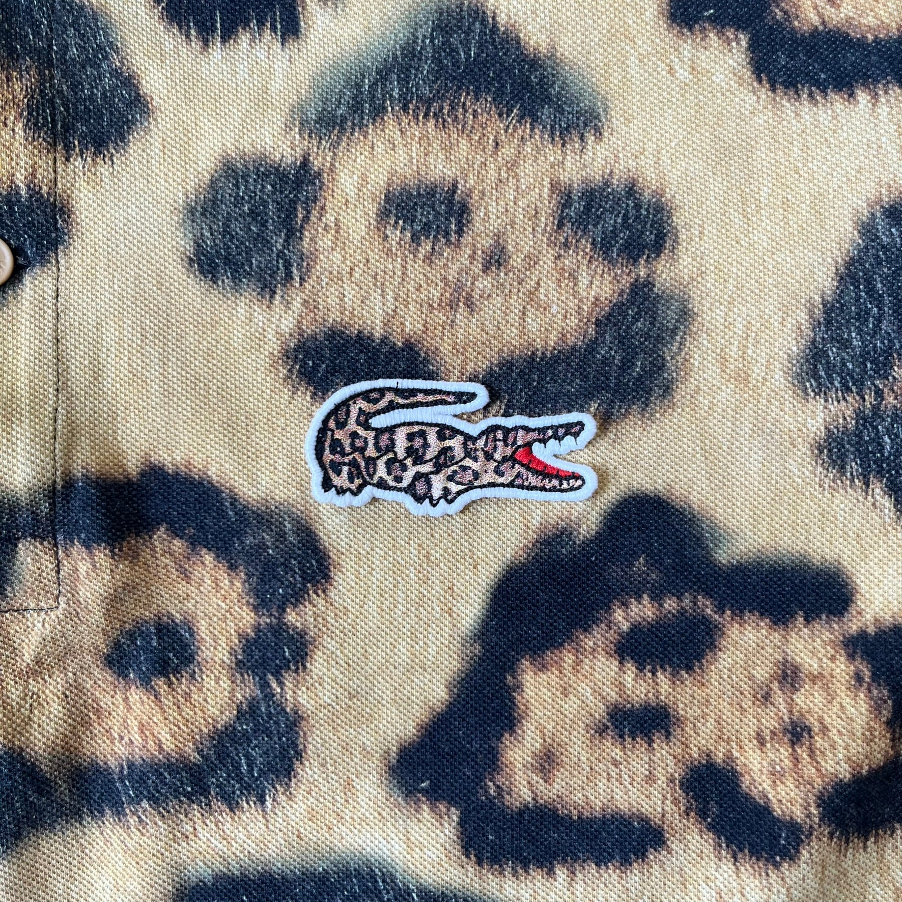 Lacoste x National Geographic AW20 Leopard Polo features an all-over beige, brown and black leopard print pattern across the fabric. The polo has beige collar and sleeve hems, with a chest patch featuring the Lacoste crocodile logo in matching pattern colors.