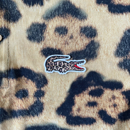 Lacoste x National Geographic AW20 Leopard Polo features an all-over beige, brown and black leopard print pattern across the fabric. The polo has beige collar and sleeve hems, with a chest patch featuring the Lacoste crocodile logo in matching pattern colors.