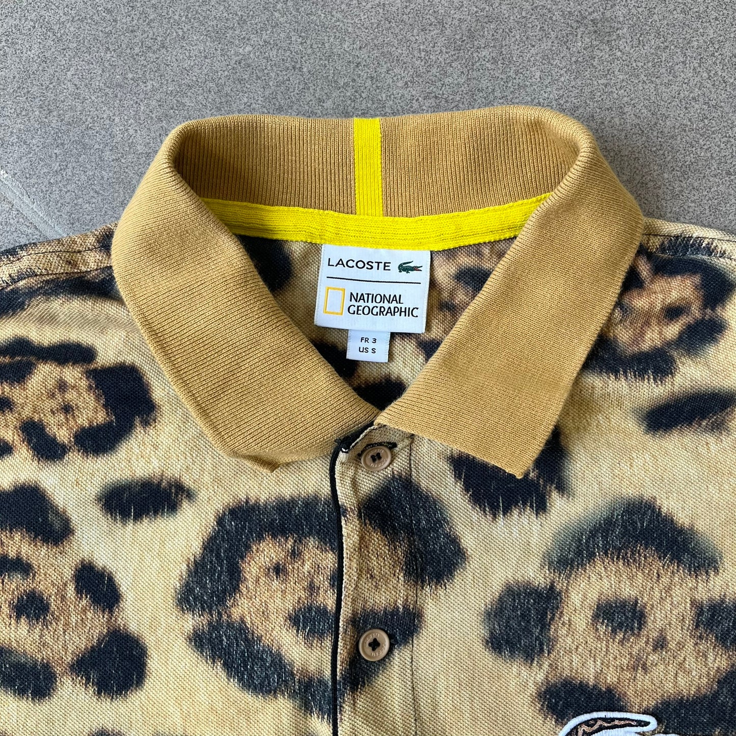 Lacoste x National Geographic AW20 Leopard Polo features an all-over beige, brown and black leopard print pattern across the fabric. The polo has beige collar and sleeve hems, with a chest patch featuring the Lacoste crocodile logo in matching pattern colors.