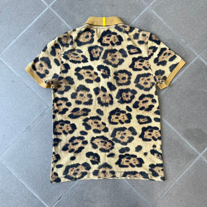 Lacoste x National Geographic AW20 Leopard Polo features an all-over beige, brown and black leopard print pattern across the fabric. The polo has beige collar and sleeve hems, with a chest patch featuring the Lacoste crocodile logo in matching pattern colors.