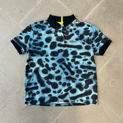 Lacoste x National Geographic AW20 Poison Dart Frog Polo features an all-over blue and black poison dart frog pattern across the fabric. The polo has black collar and sleeve hems, with a chest patch featuring the Lacoste crocodile logo in matching pattern colors.
