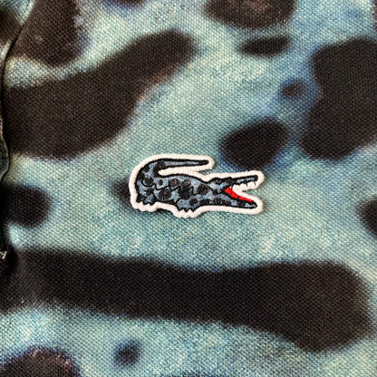 Lacoste x National Geographic AW20 Poison Dart Frog Polo features an all-over blue and black poison dart frog pattern across the fabric. The polo has black collar and sleeve hems, with a chest patch featuring the Lacoste crocodile logo in matching pattern colors.
