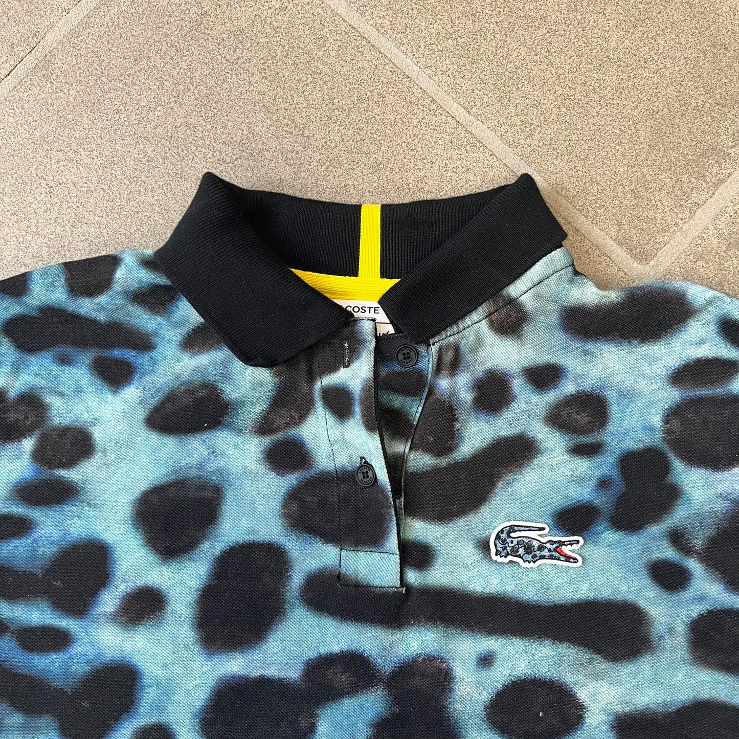 Lacoste x National Geographic AW20 Poison Dart Frog Polo features an all-over blue and black poison dart frog pattern across the fabric. The polo has black collar and sleeve hems, with a chest patch featuring the Lacoste crocodile logo in matching pattern colors.
