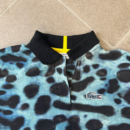 Lacoste x National Geographic AW20 Poison Dart Frog Polo features an all-over blue and black poison dart frog pattern across the fabric. The polo has black collar and sleeve hems, with a chest patch featuring the Lacoste crocodile logo in matching pattern colors.
