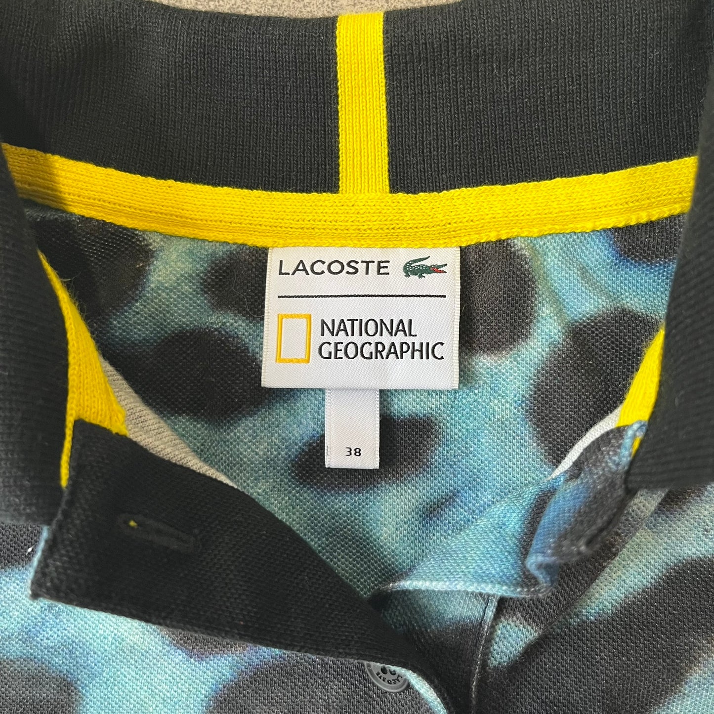 Lacoste x National Geographic AW20 Poison Dart Frog Polo features an all-over blue and black poison dart frog pattern across the fabric. The polo has black collar and sleeve hems, with a chest patch featuring the Lacoste crocodile logo in matching pattern colors.
