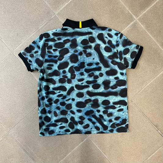 Lacoste x National Geographic AW20 Poison Dart Frog Polo features an all-over blue and black poison dart frog pattern across the fabric. The polo has black collar and sleeve hems, with a chest patch featuring the Lacoste crocodile logo in matching pattern colors.
