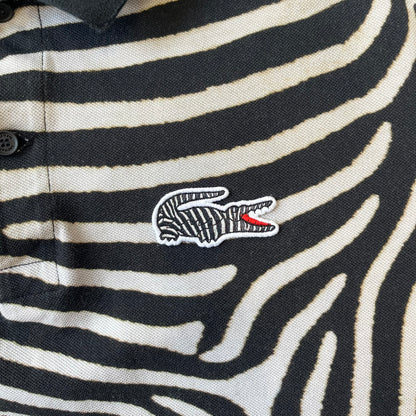  Lacoste x National Geographic AW20 Zebra Polo features an all-over white and black zebra print pattern across the fabric. The polo has black collar and sleeve hems, with a chest patch featuring the Lacoste crocodile logo in matching pattern colors.