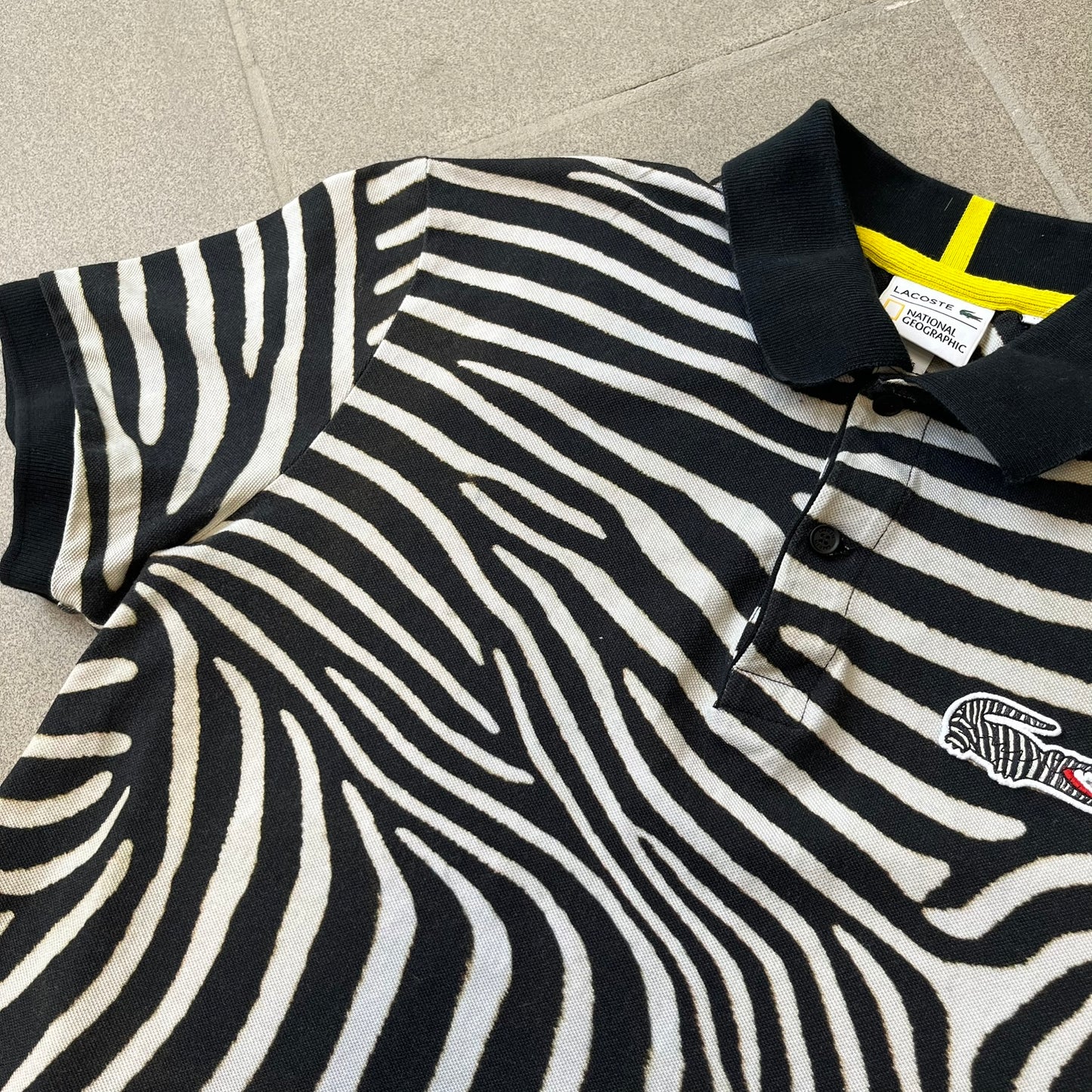  Lacoste x National Geographic AW20 Zebra Polo features an all-over white and black zebra print pattern across the fabric. The polo has black collar and sleeve hems, with a chest patch featuring the Lacoste crocodile logo in matching pattern colors.