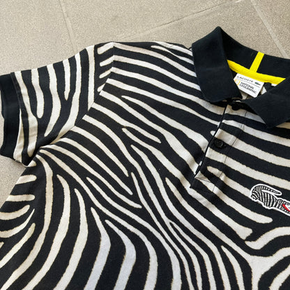  Lacoste x National Geographic AW20 Zebra Polo features an all-over white and black zebra print pattern across the fabric. The polo has black collar and sleeve hems, with a chest patch featuring the Lacoste crocodile logo in matching pattern colors.