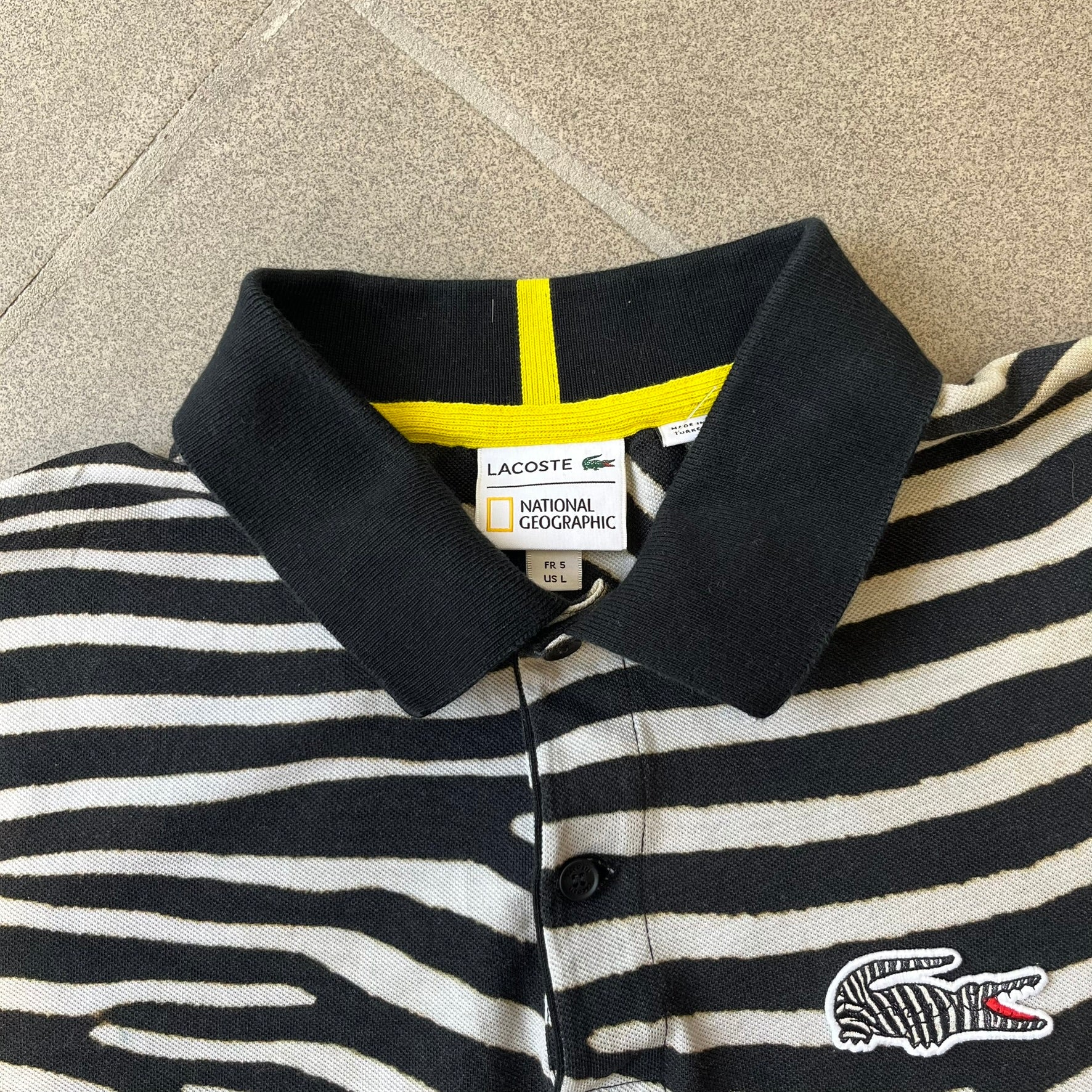  Lacoste x National Geographic AW20 Zebra Polo features an all-over white and black zebra print pattern across the fabric. The polo has black collar and sleeve hems, with a chest patch featuring the Lacoste crocodile logo in matching pattern colors.