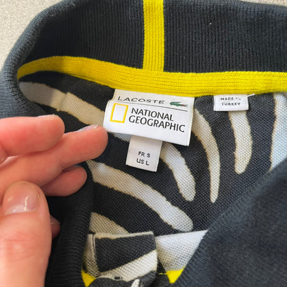  Lacoste x National Geographic AW20 Zebra Polo features an all-over white and black zebra print pattern across the fabric. The polo has black collar and sleeve hems, with a chest patch featuring the Lacoste crocodile logo in matching pattern colors.