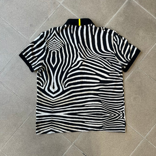  Lacoste x National Geographic AW20 Zebra Polo features an all-over white and black zebra print pattern across the fabric. The polo has black collar and sleeve hems, with a chest patch featuring the Lacoste crocodile logo in matching pattern colors.