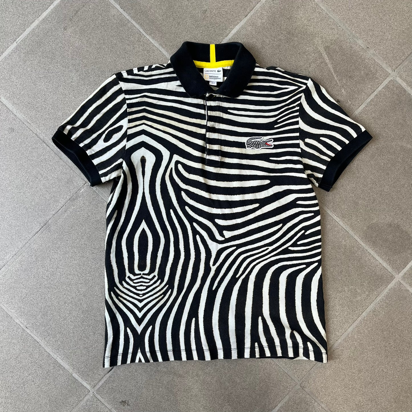 Lacoste x National Geographic AW20 Zebra Polo features an all-over white and black zebra print pattern across the fabric. The polo has black collar and sleeve hems, with a chest patch featuring the Lacoste crocodile logo in matching pattern colors.
