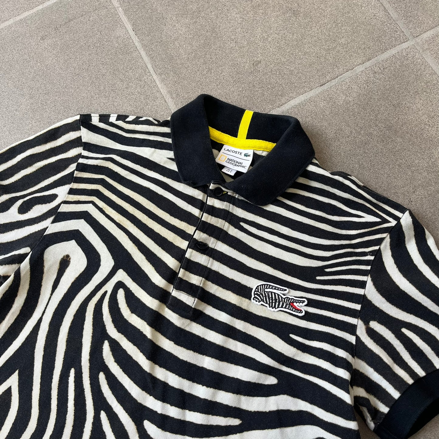 Lacoste x National Geographic AW20 Zebra Polo features an all-over white and black zebra print pattern across the fabric. The polo has black collar and sleeve hems, with a chest patch featuring the Lacoste crocodile logo in matching pattern colors.