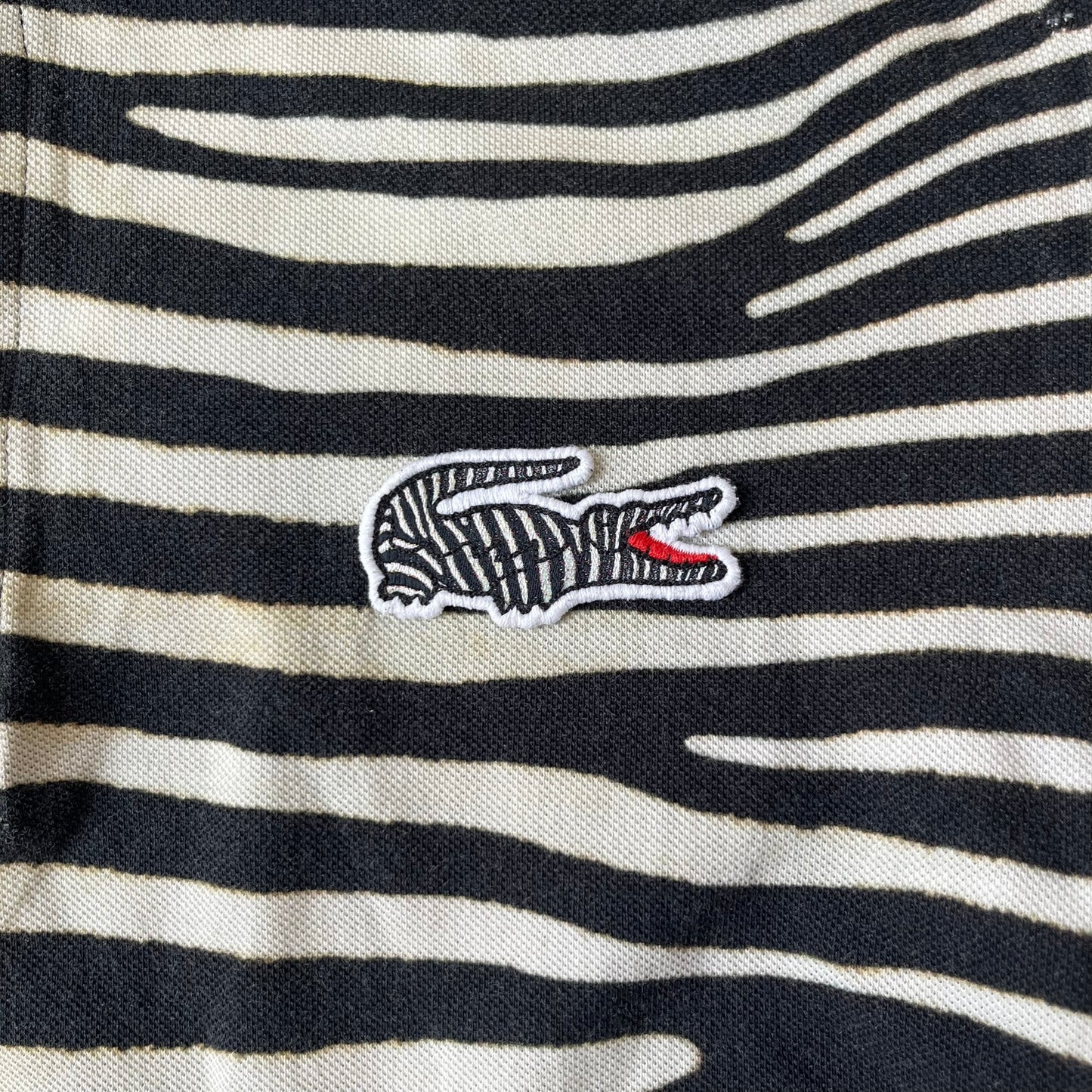 Lacoste x National Geographic AW20 Zebra Polo features an all-over white and black zebra print pattern across the fabric. The polo has black collar and sleeve hems, with a chest patch featuring the Lacoste crocodile logo in matching pattern colors.
