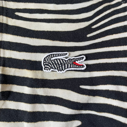 Lacoste x National Geographic AW20 Zebra Polo features an all-over white and black zebra print pattern across the fabric. The polo has black collar and sleeve hems, with a chest patch featuring the Lacoste crocodile logo in matching pattern colors.