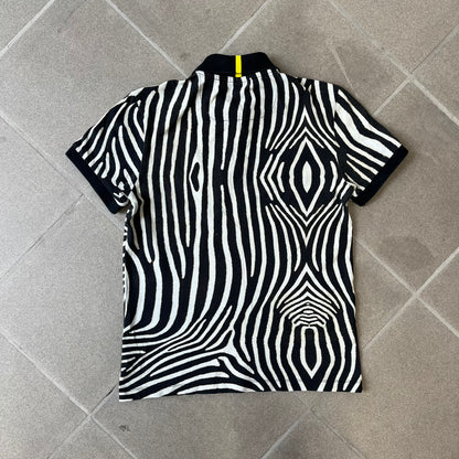 Lacoste x National Geographic AW20 Zebra Polo features an all-over white and black zebra print pattern across the fabric. The polo has black collar and sleeve hems, with a chest patch featuring the Lacoste crocodile logo in matching pattern colors.