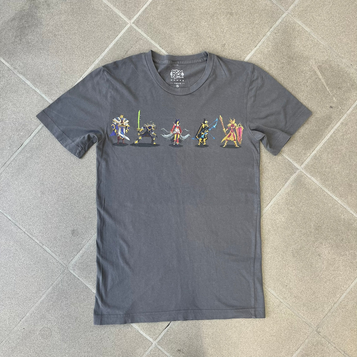 League of Legends 2013 Pixel Team Gray Tee is a gray T-shirt featuring a full five-champion team (Garen, Master Yi, Ahri, Ashe, and Kayle) rendered in pixel art style on the front. The upper back shows League main Logo. Licensed 2013 neck tag with the old Riot Games logo.