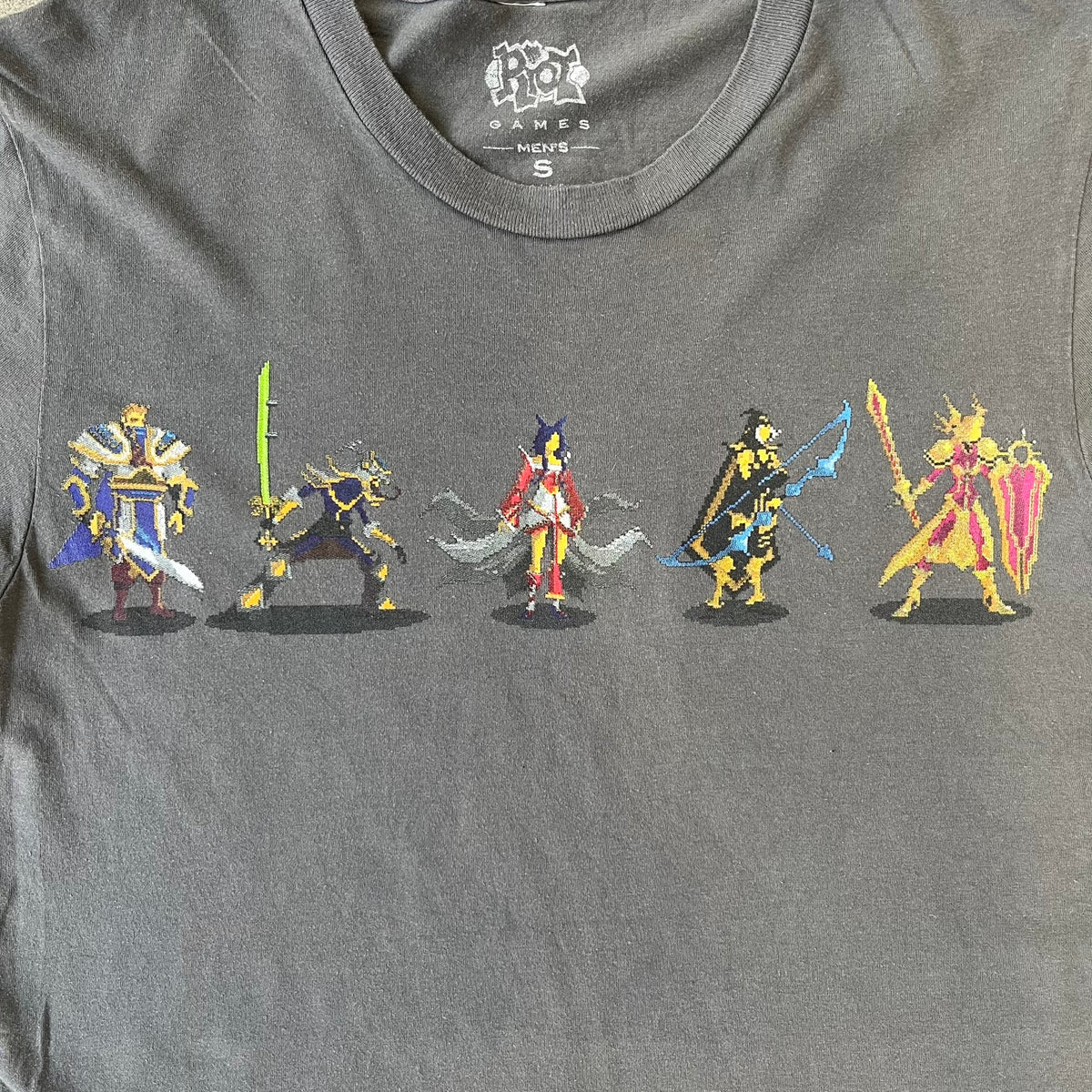 League of Legends 2013 Pixel Team Gray Tee is a gray T-shirt featuring a full five-champion team (Garen, Master Yi, Ahri, Ashe, and Kayle) rendered in pixel art style on the front. The upper back shows League main Logo. Licensed 2013 neck tag with the old Riot Games logo.