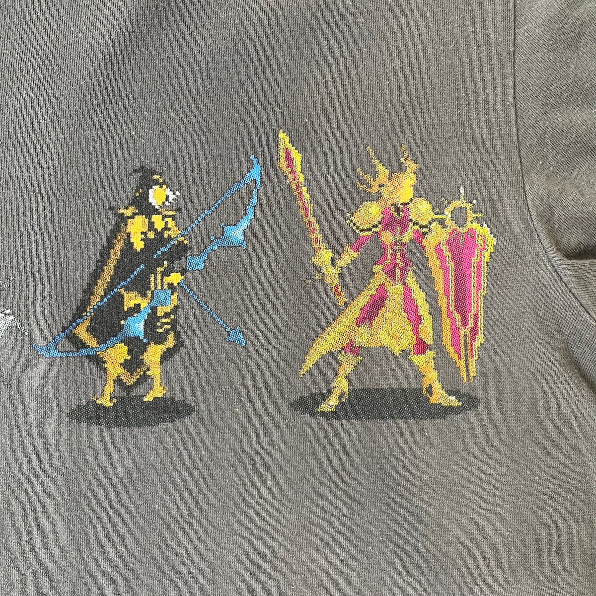 League of Legends 2013 Pixel Team Gray Tee is a gray T-shirt featuring a full five-champion team (Garen, Master Yi, Ahri, Ashe, and Kayle) rendered in pixel art style on the front. The upper back shows League main Logo. Licensed 2013 neck tag with the old Riot Games logo.