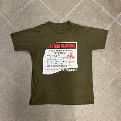 Left 4 Dead 2009 Infection Warning Tee features a khaki T-shirt with a distressed poster-style graphic on the front. The graphic has red “Infection Warning” text, instructions on what to do when infected, an emergency hotline number, and scribbled details. A Left 4 Dead logo and small 2009 Valve licensing appear underneath. The upper back features a Left 4 Dead logo with a blood splatter graphic.