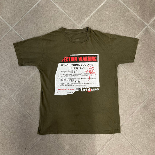 Left 4 Dead 2009 Infection Warning Tee features a khaki T-shirt with a distressed poster-style graphic on the front. The graphic has red “Infection Warning” text, instructions on what to do when infected, an emergency hotline number, and scribbled details. A Left 4 Dead logo and small 2009 Valve licensing appear underneath. The upper back features a Left 4 Dead logo with a blood splatter graphic.