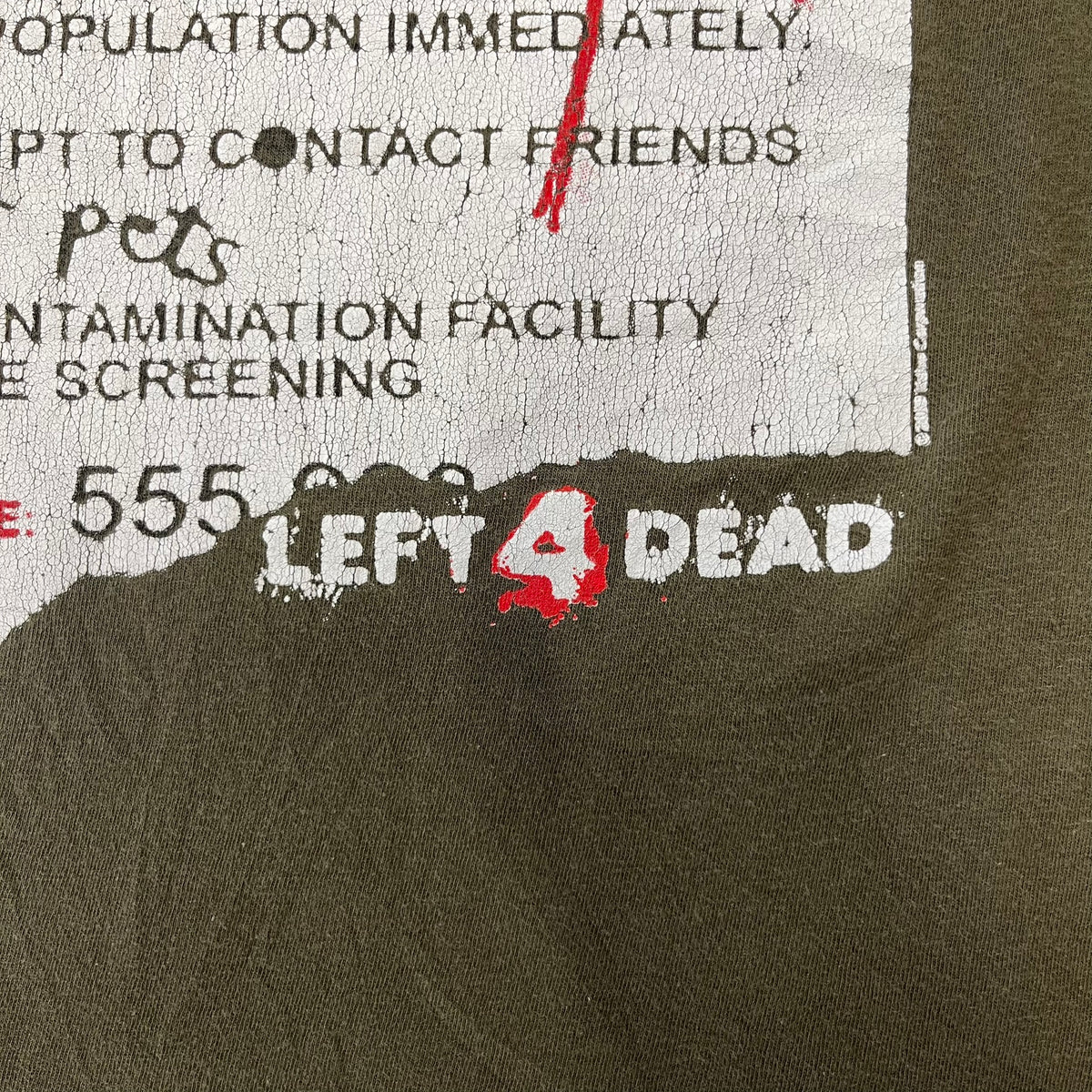 Left 4 Dead 2009 Infection Warning Tee features a khaki T-shirt with a distressed poster-style graphic on the front. The graphic has red “Infection Warning” text, instructions on what to do when infected, an emergency hotline number, and scribbled details. A Left 4 Dead logo and small 2009 Valve licensing appear underneath. The upper back features a Left 4 Dead logo with a blood splatter graphic.