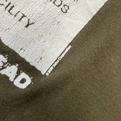 Left 4 Dead 2009 Infection Warning Tee features a khaki T-shirt with a distressed poster-style graphic on the front. The graphic has red “Infection Warning” text, instructions on what to do when infected, an emergency hotline number, and scribbled details. A Left 4 Dead logo and small 2009 Valve licensing appear underneath. The upper back features a Left 4 Dead logo with a blood splatter graphic.