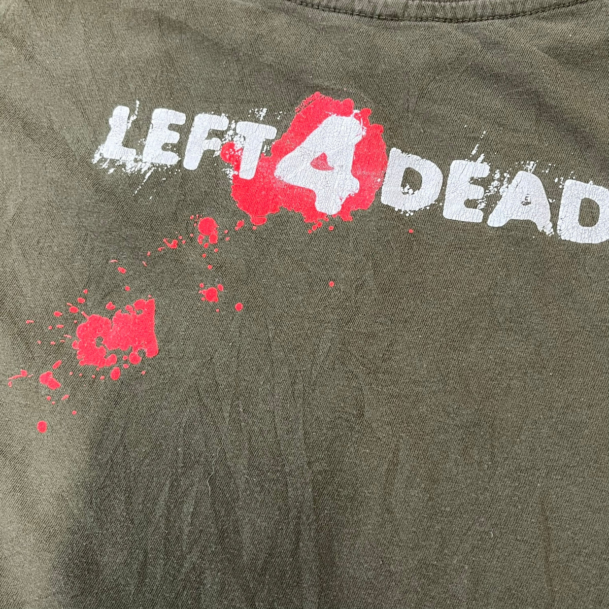 Left 4 Dead 2009 Infection Warning Tee features a khaki T-shirt with a distressed poster-style graphic on the front. The graphic has red “Infection Warning” text, instructions on what to do when infected, an emergency hotline number, and scribbled details. A Left 4 Dead logo and small 2009 Valve licensing appear underneath. The upper back features a Left 4 Dead logo with a blood splatter graphic.