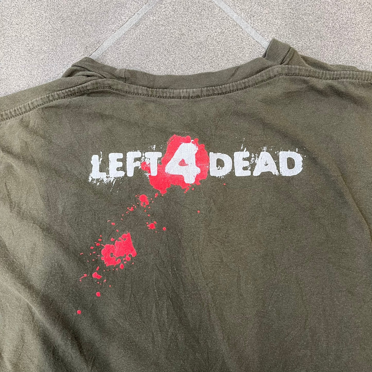 Left 4 Dead 2009 Infection Warning Tee features a khaki T-shirt with a distressed poster-style graphic on the front. The graphic has red “Infection Warning” text, instructions on what to do when infected, an emergency hotline number, and scribbled details. A Left 4 Dead logo and small 2009 Valve licensing appear underneath. The upper back features a Left 4 Dead logo with a blood splatter graphic.